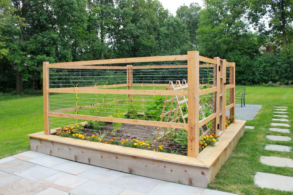 Raised vegetable or flower bed with wood rail, stone patio, lawn stepping path, and wooded backyard.
